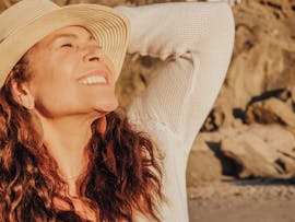 A joyful woman with a hat basks in the sunlight by the seaside, symbolizing relaxation and happiness.
