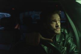 Bearded man in a leather jacket driving a car at night, looking thoughtfully out the window.
