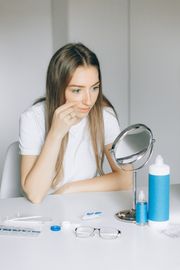 Young woman in white shirt applying contact lens with mirror on table.